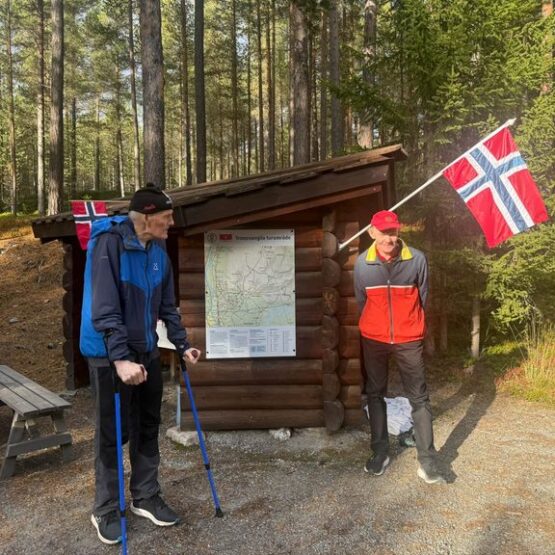 To menn med norske flagg og turstaver står utenfor en tømmergapahuk med kart over Tronsvanglia turområde.