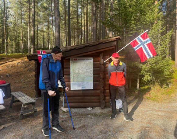 To menn med norske flagg og turstaver står utenfor en tømmergapahuk med kart over Tronsvanglia turområde.