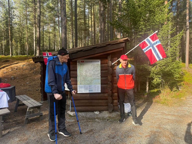 To menn med norske flagg og turstaver står utenfor en tømmergapahuk med kart over Tronsvanglia turområde.