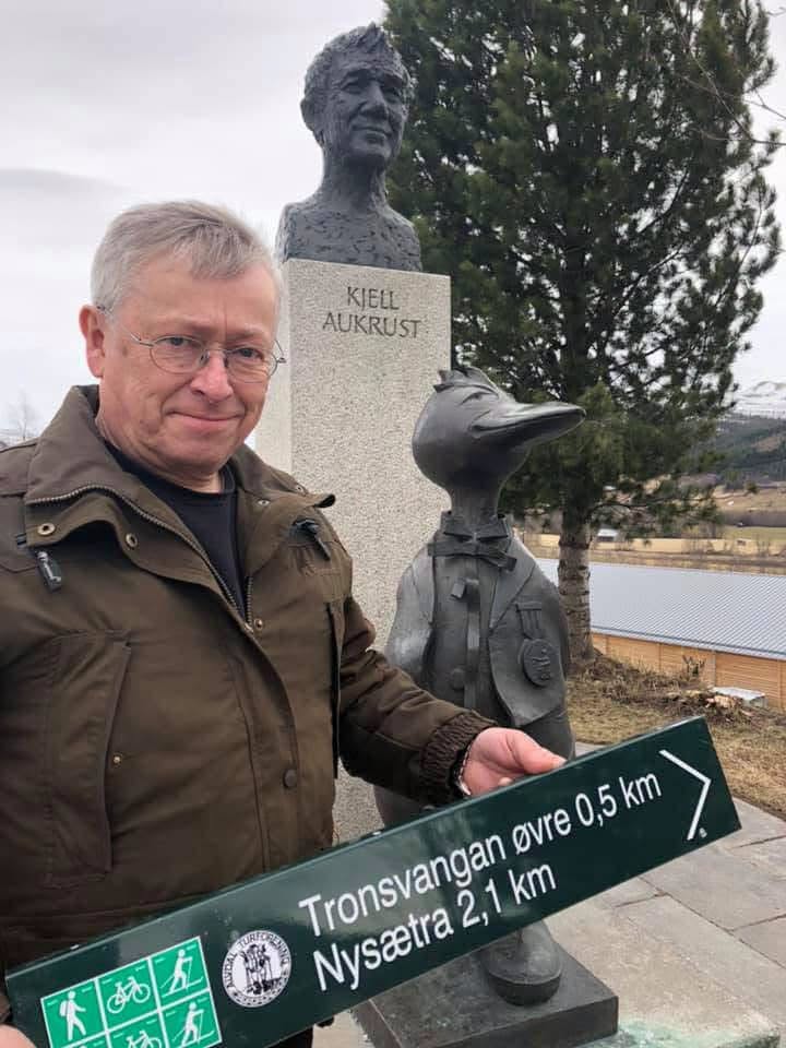 En mann står ved statuen av Kjell Aukrust og holder et skilt som viser vei til Tronsvangan øvre og Nysætra.