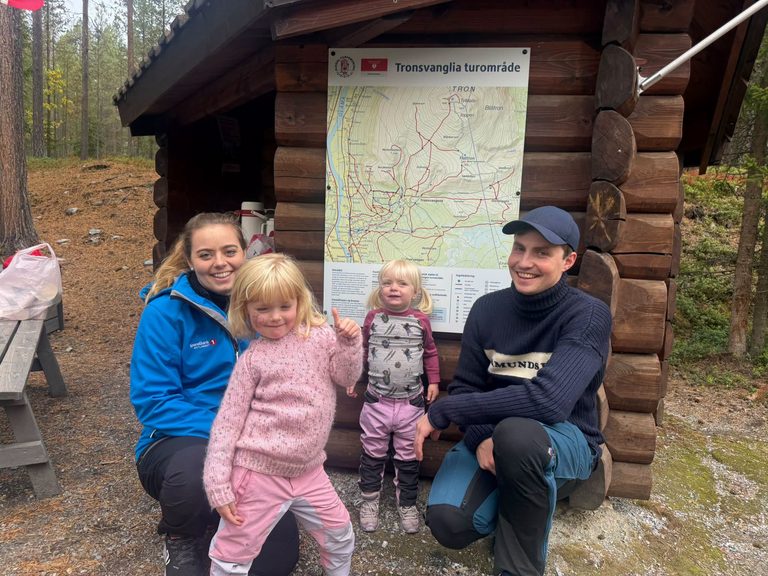 En kvinne og en mann sitter på huk med to jenter foran kartplakaten for Tronsvanglia turområde.