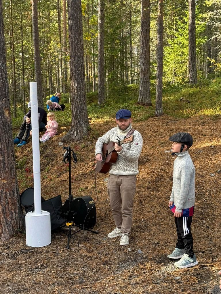 En mann spiller gitar og en gutt synger, mens andre barn og voksne sitter og lytter i skogen.