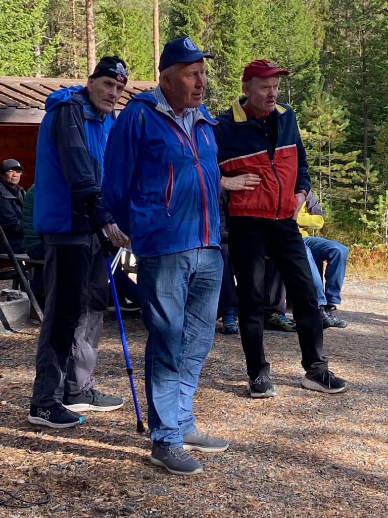 Tre eldre menn i turjakker står samlet utendørs, en av dem støtter seg til en turstav.